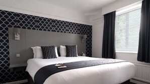 Une chambre d’hôtel avec un grand lit, une tête de lit ornée d’un motif géométrique, une table de chevet, une fenêtre avec des rideaux et un lampadaire fixé au mur.