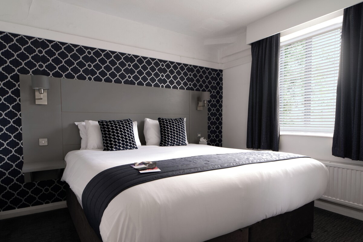Une chambre d’hôtel avec un grand lit, une tête de lit ornée d’un motif géométrique, une table de chevet, une fenêtre avec des rideaux et un lampadaire fixé au mur.