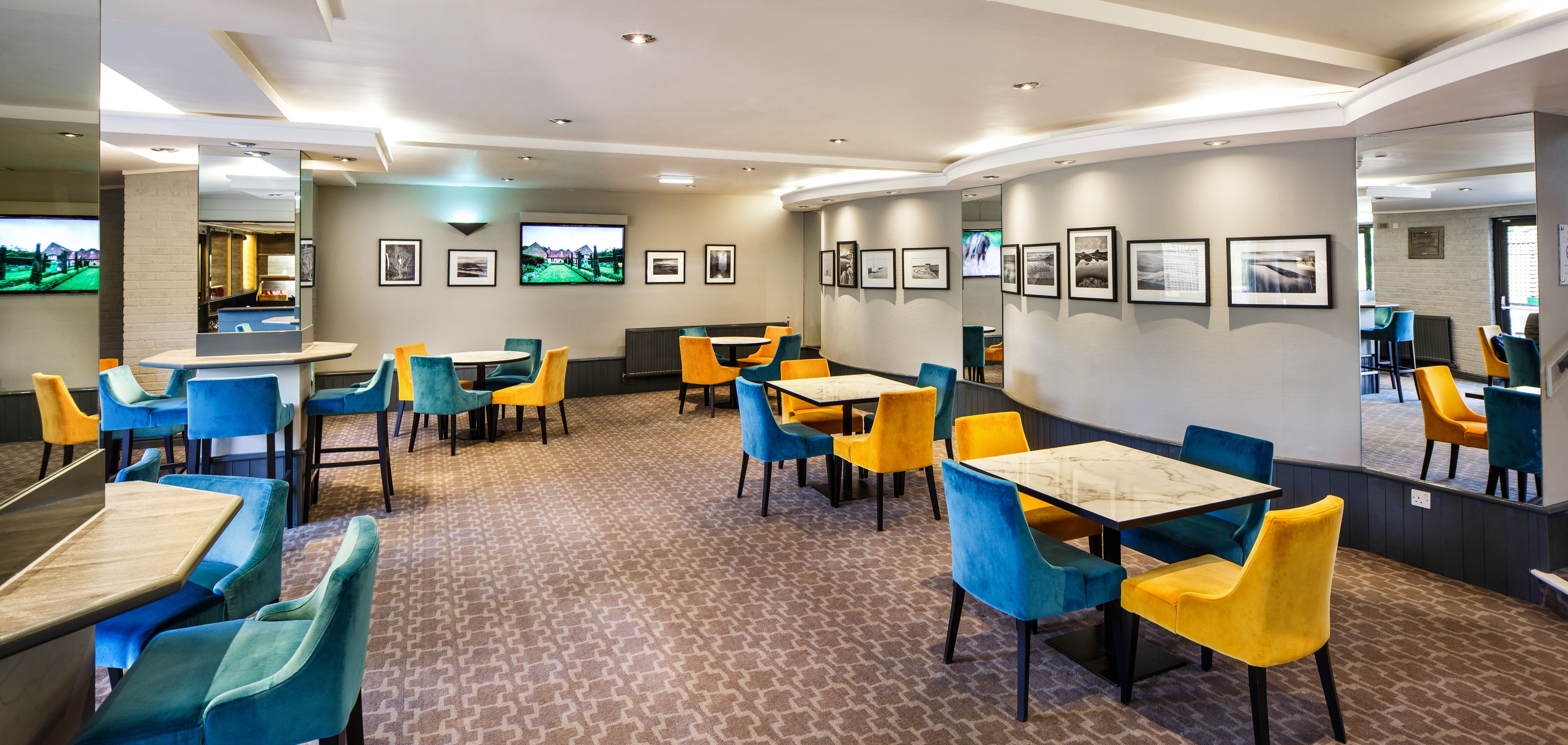 A hotel dining area with multiple tables and chairs, a television screen, and framed pictures on the walls.