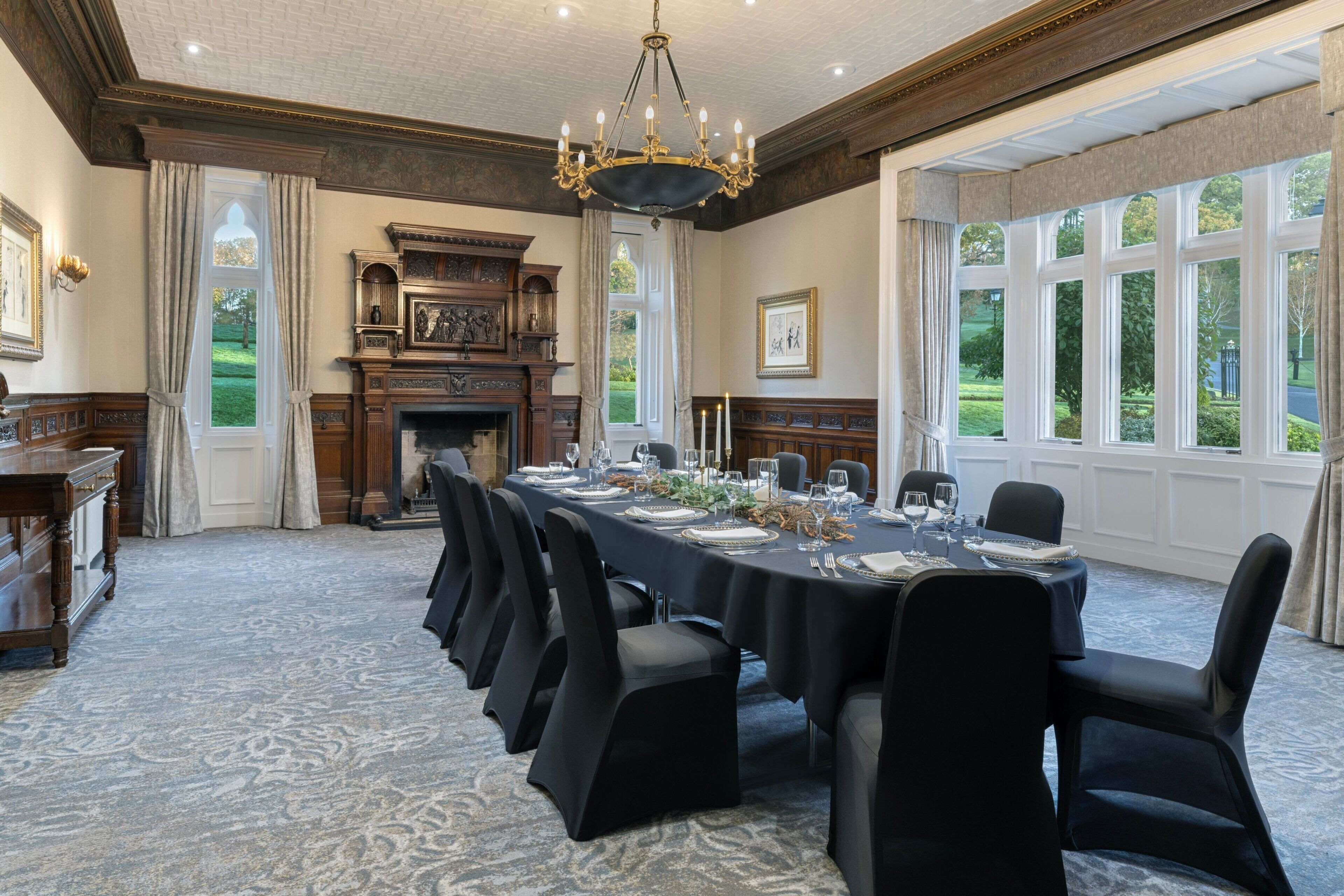 A formal dining room with a long table set for a meal, flanked by chairs, a fireplace, and large windows offering a view of the outdoors.