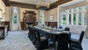 A formal dining room with a long table set for a meal, flanked by chairs, a fireplace, and large windows offering a view of the outdoors.
