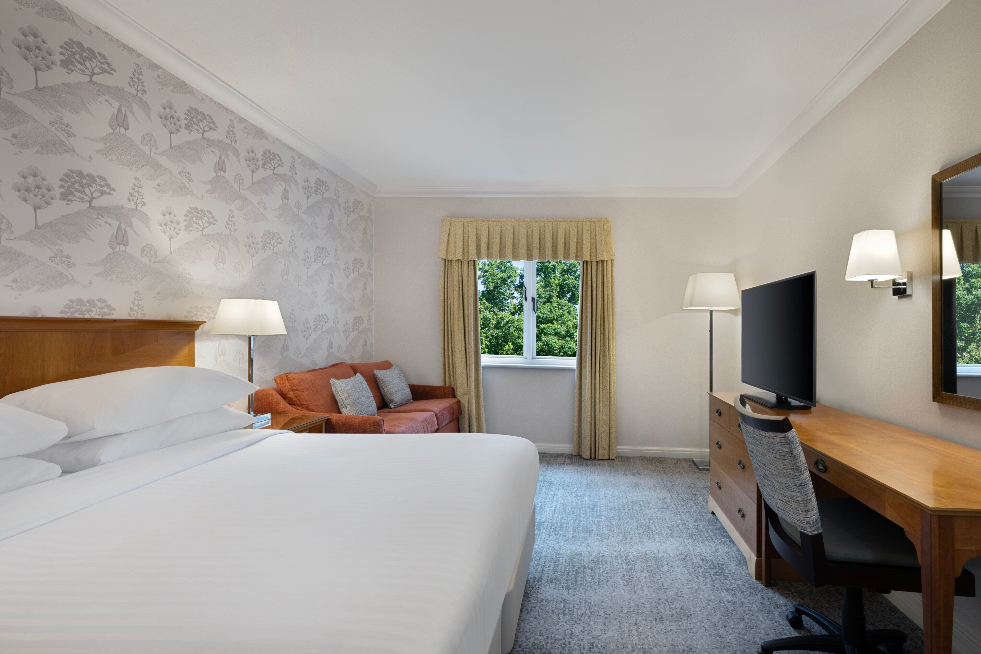 A hotel room with a large bed, a desk with a computer, a TV, and a window with curtains.