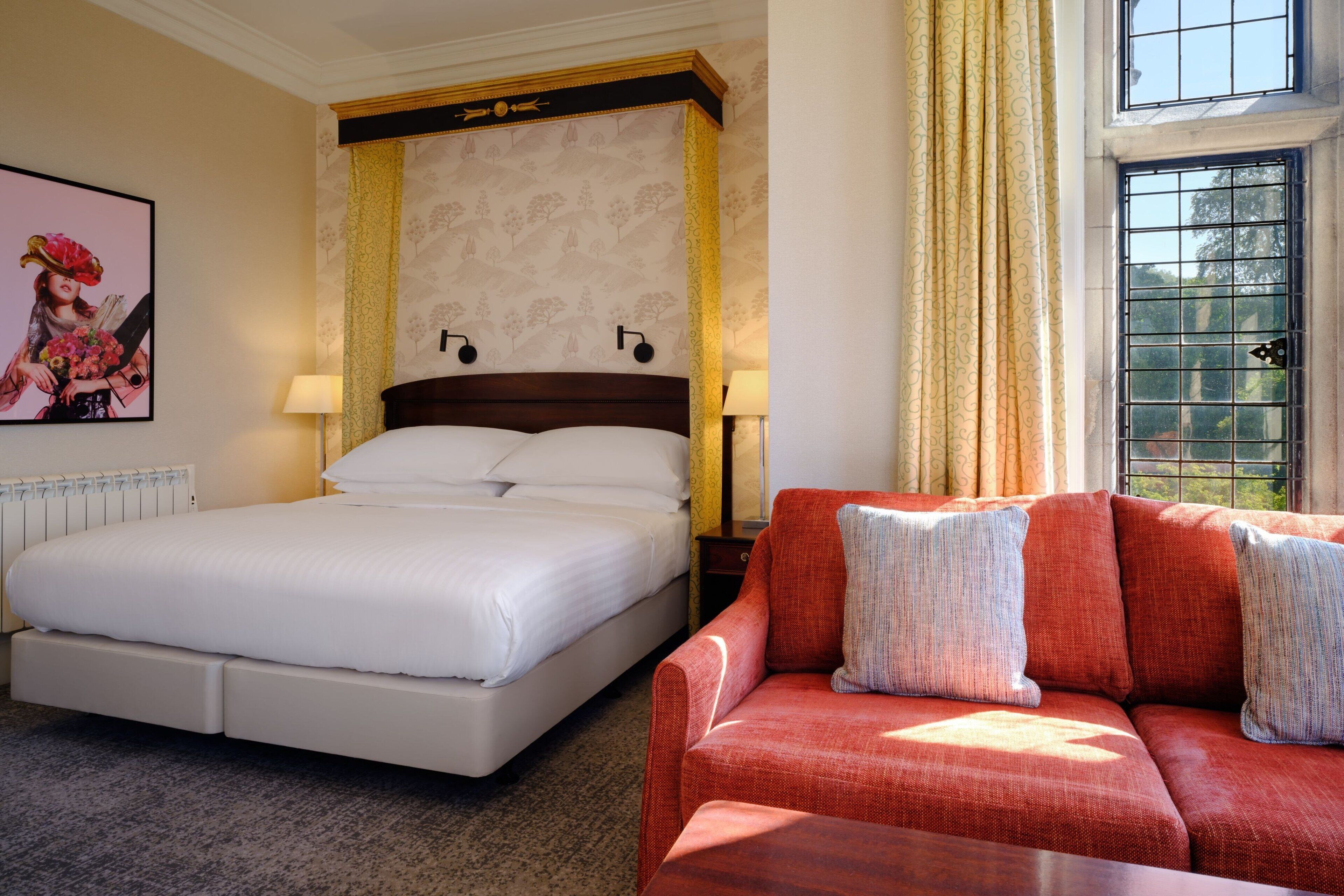 A hotel room with a large bed, a red sofa, a nightstand, a lamp, a framed picture on the wall, and a window with curtains.