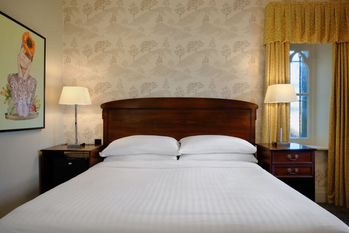 A neatly made bed with white linens, two bedside tables with lamps, a wooden headboard, and a window with curtains.