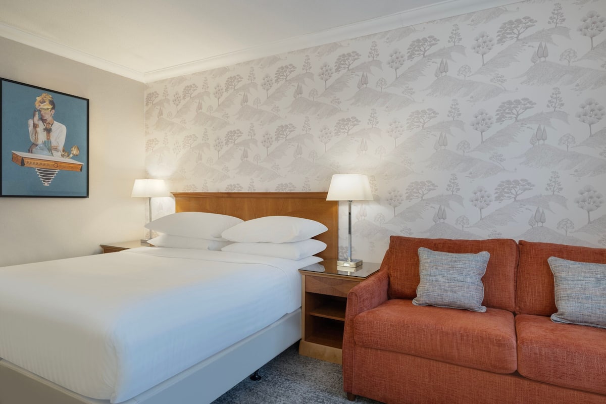 A hotel room with a bed, a nightstand, a lamp, a sofa, and a wall with a tree pattern.