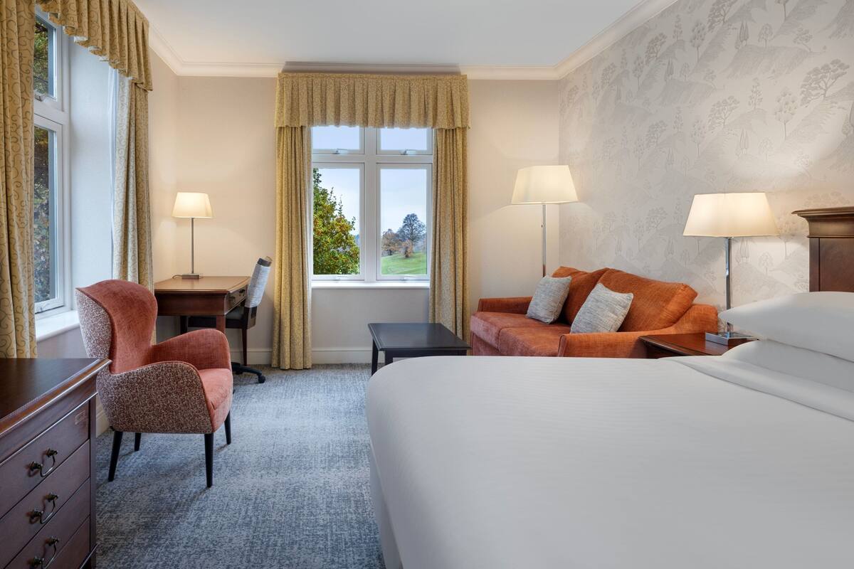 A hotel room with a bed, two chairs, a desk, and a view of a green landscape through the window.
