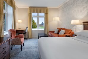 A hotel room with a bed, two chairs, a desk, and a view of a green landscape through the window. - Delta Hotels by Marriott Breadsall Priory Country Club (Derby)