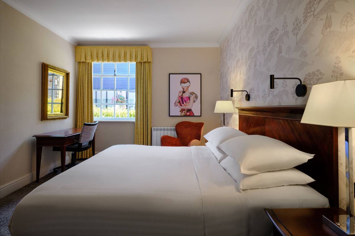 A hotel room with a large bed, a desk, a chair, a window with curtains, a wall-mounted lamp, and a framed picture.