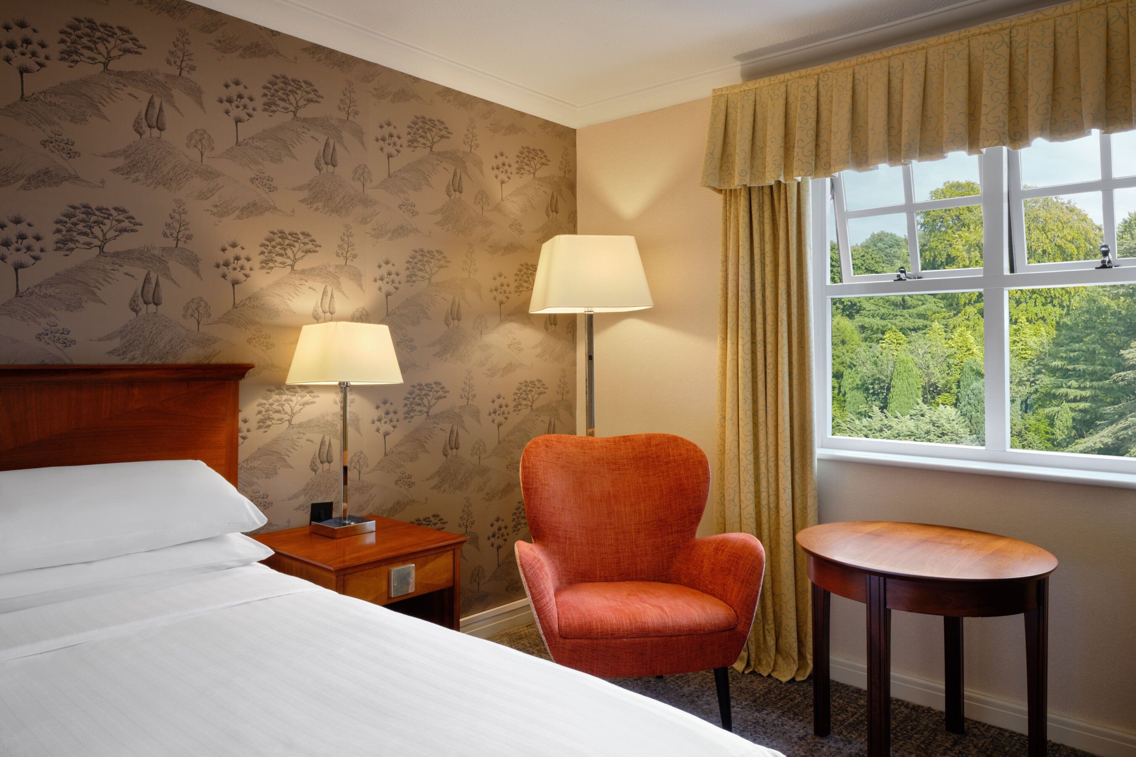 Habitación de hotel con cama, dos lámparas de noche, una silla, una mesita y una ventana con vistas a zonas verdes.