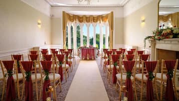 Un espacio para eventos formales con filas de sillas, un pasillo alfombrado y un arreglo floral en la parte delantera.