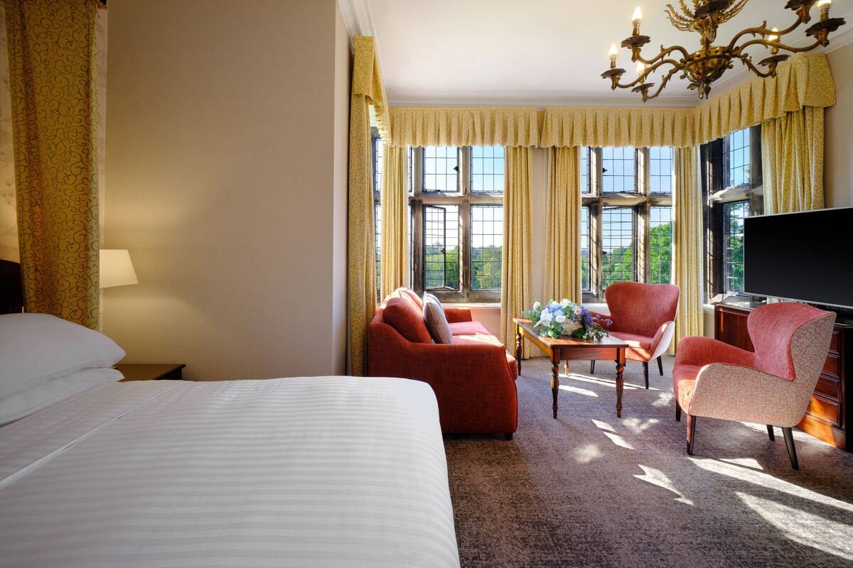A hotel room with a large bed, a sofa, a chair, a table with flowers, and a TV.