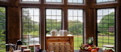 A breakfast setup with tea, coffee, and fruit in a room with large windows overlooking a garden.
