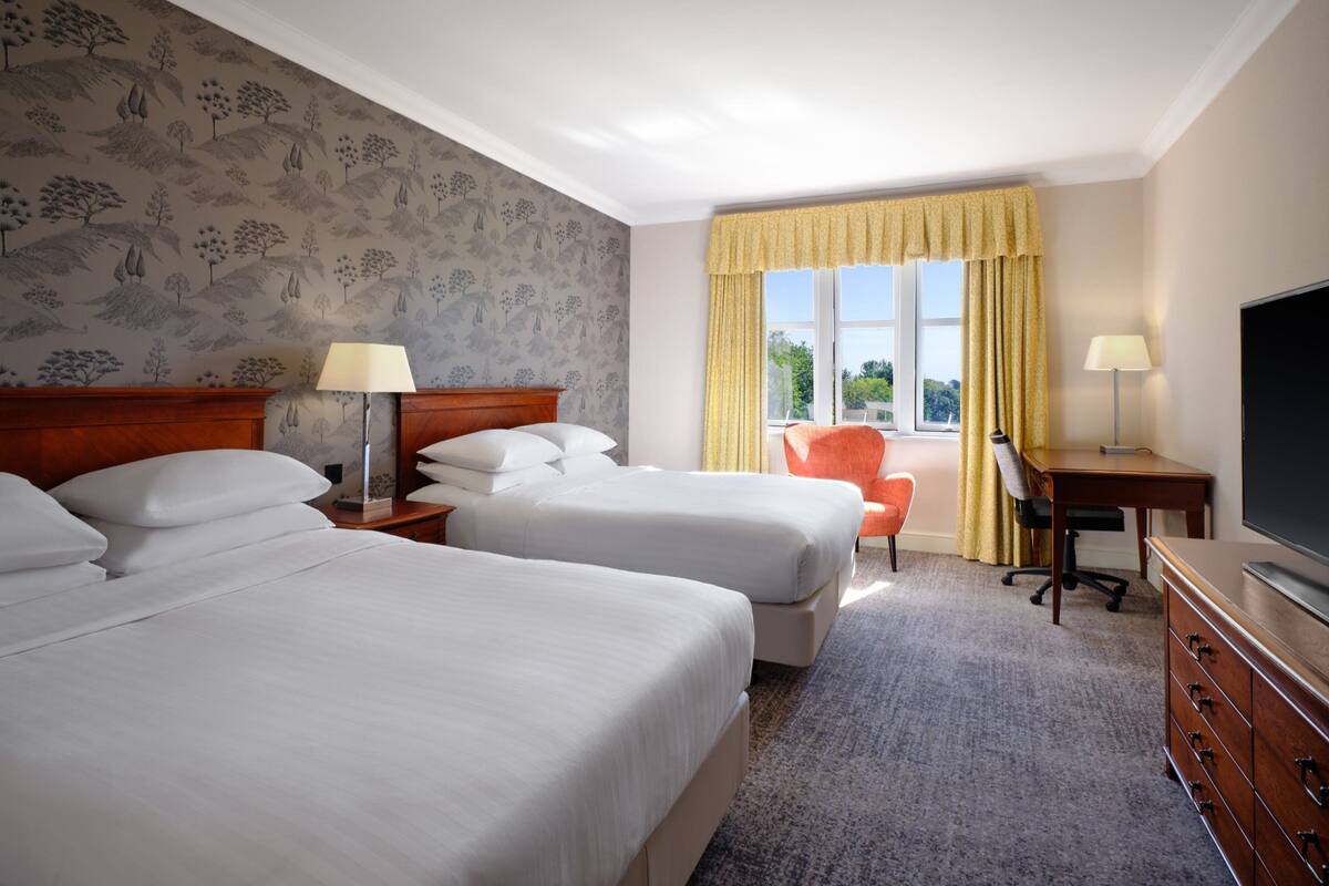 A hotel room with two beds, a desk, a chair, a TV, and a window with curtains.