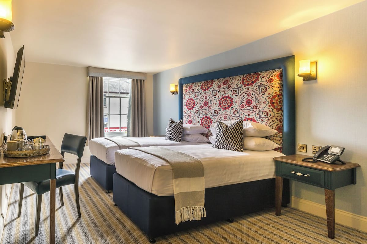 Una habitación de hotel con cama, mesitas de noche, un escritorio con silla, ventana con cortinas y cabecera estampada.