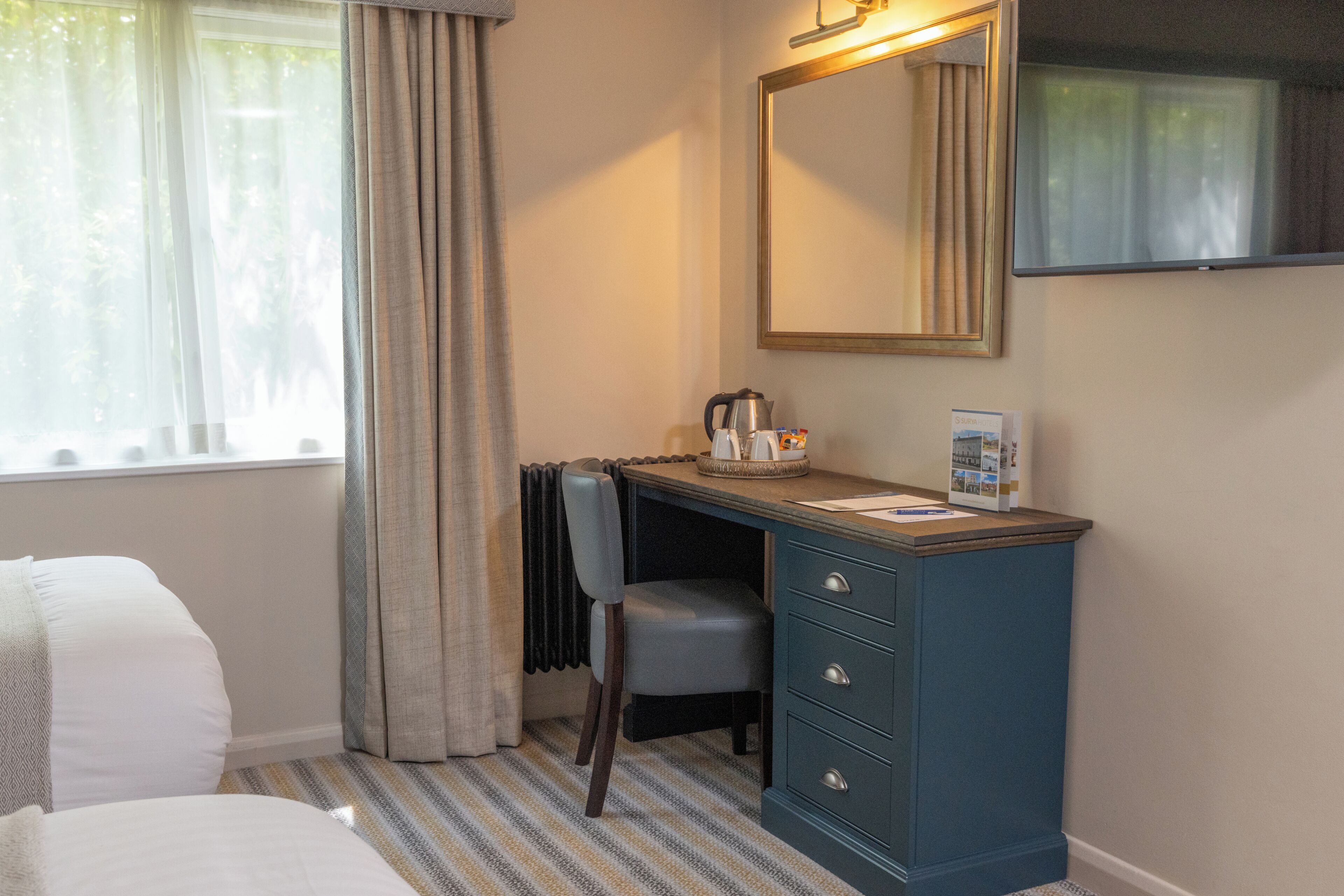 A hotel room with a bed, a desk with a chair, a radiator, a mirror, and a window with curtains.