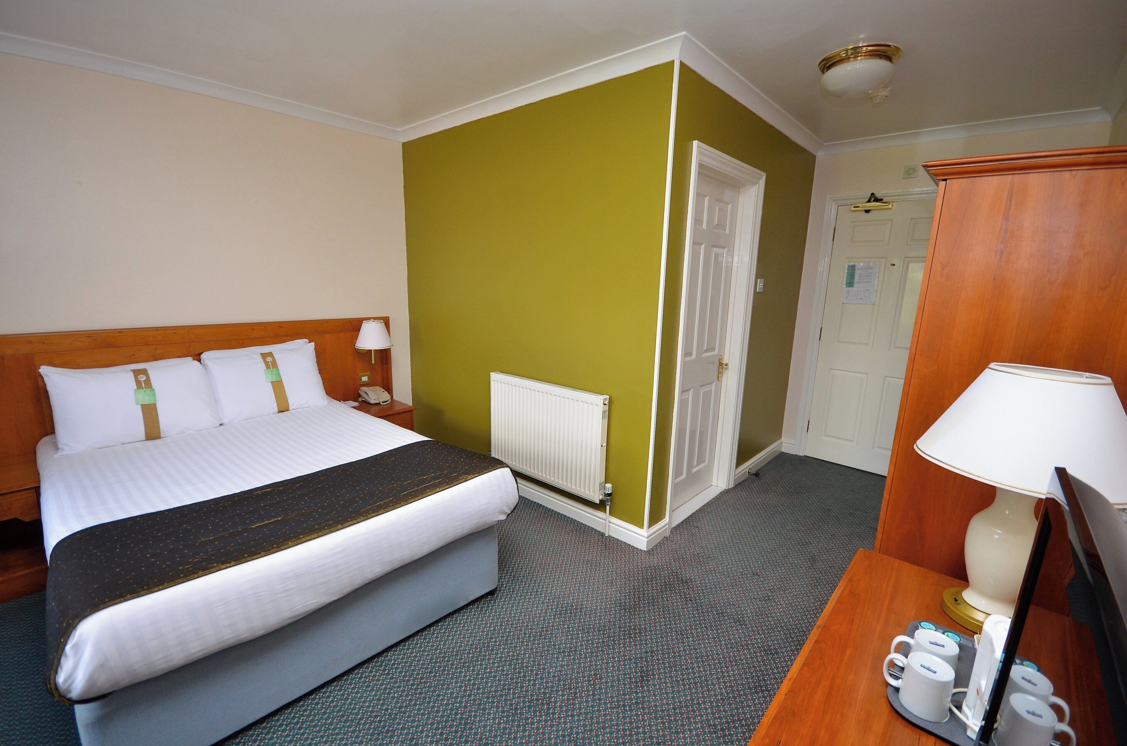 A hotel room with a bed, a desk with a lamp, a radiator, and a door.