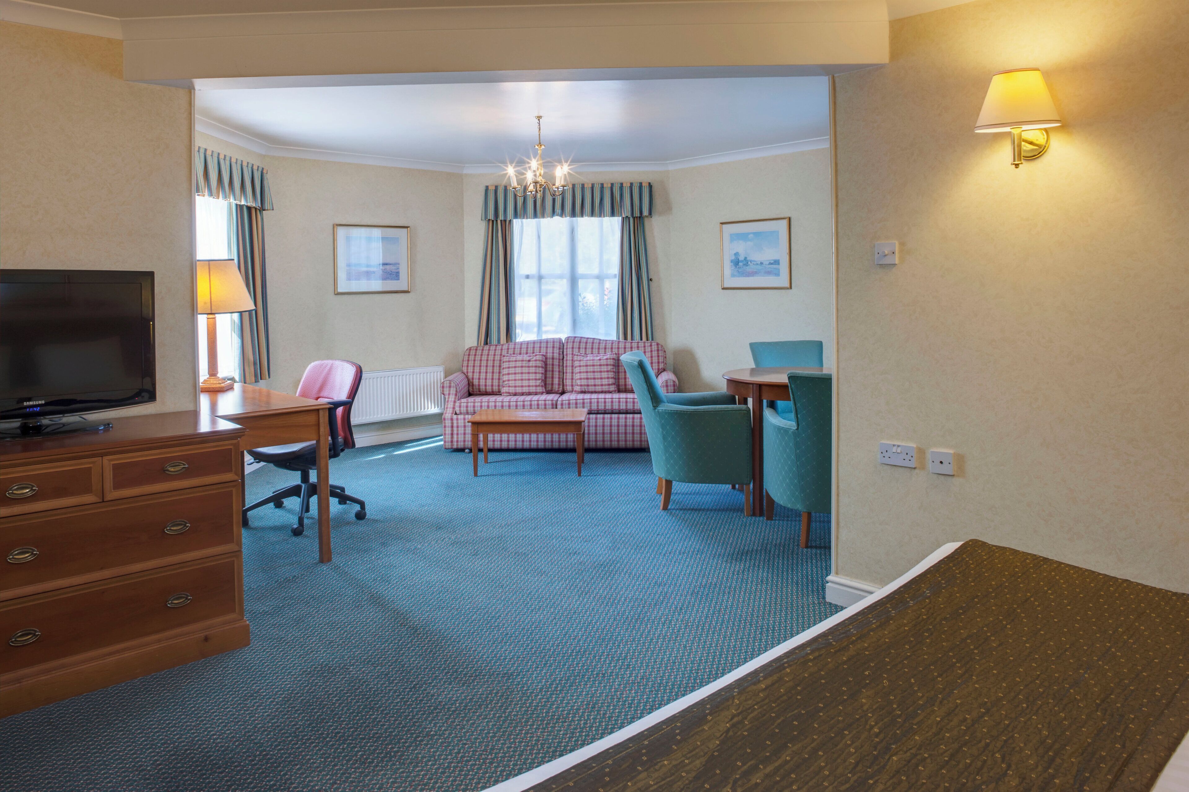 A hotel room with a television, a sofa, a chair, a desk, and a lamp.