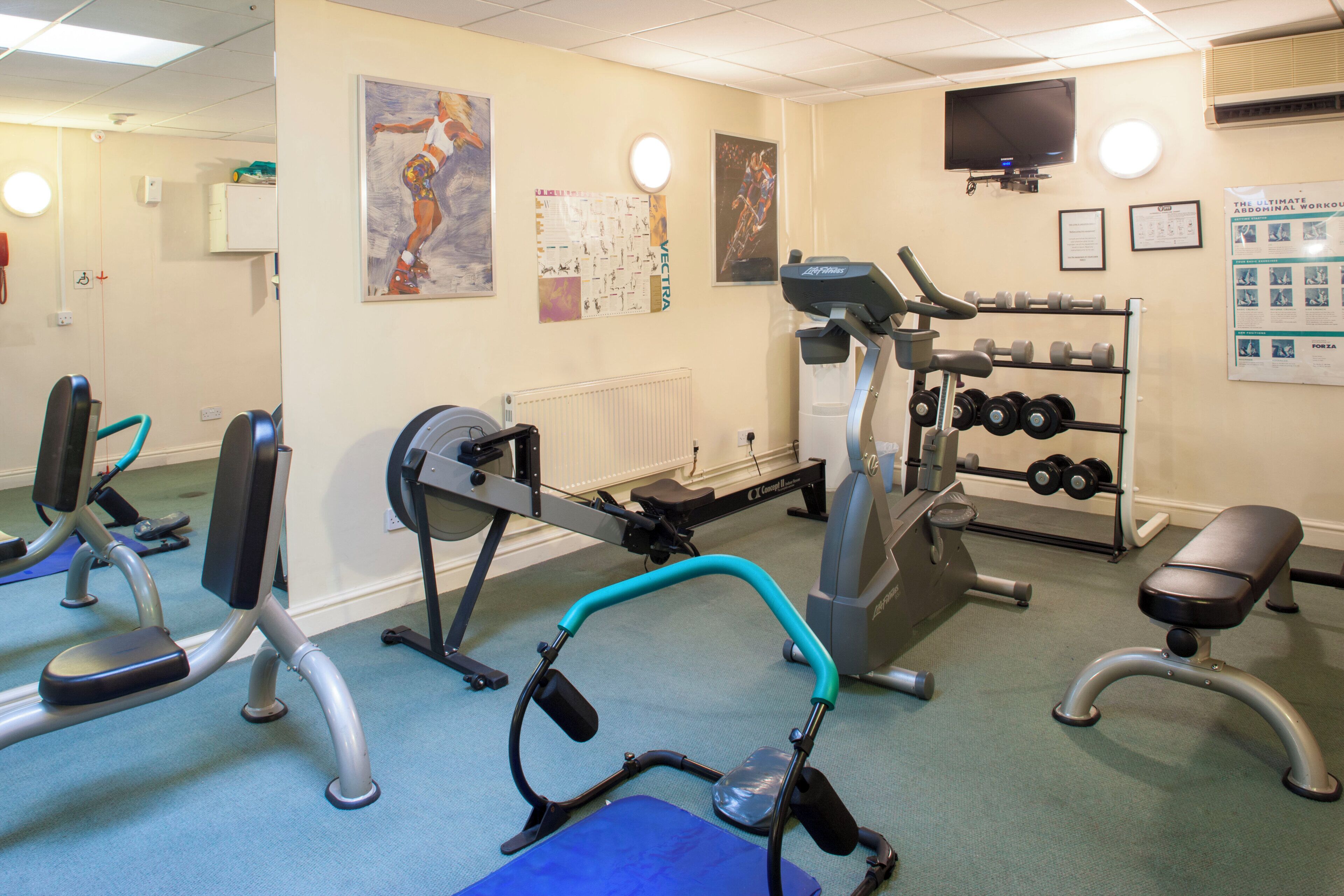 Une salle de sport équipée de matériel d’exercice, d’un téléviseur et de panneaux d’information accrochés aux murs.