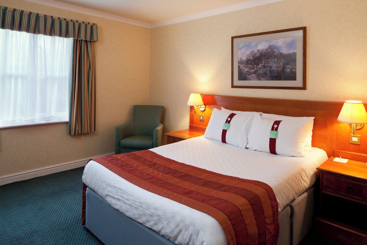 A hotel room with a large bed, two bedside lamps, a chair, a painting on the wall, and a window with curtains.