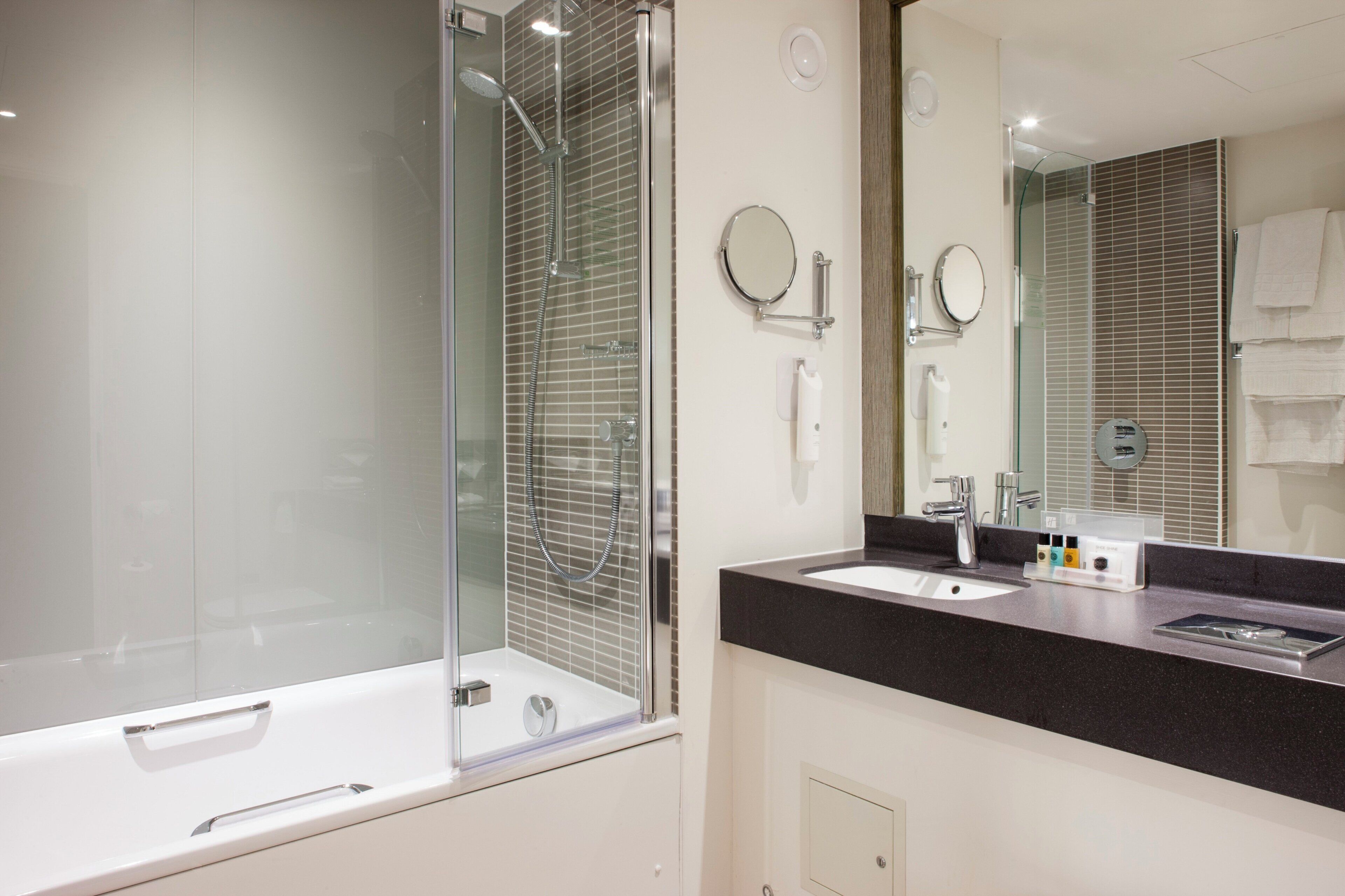 Un baño moderno con ducha de vidrio, un espejo grande y una encimera negra con artículos de aseo.