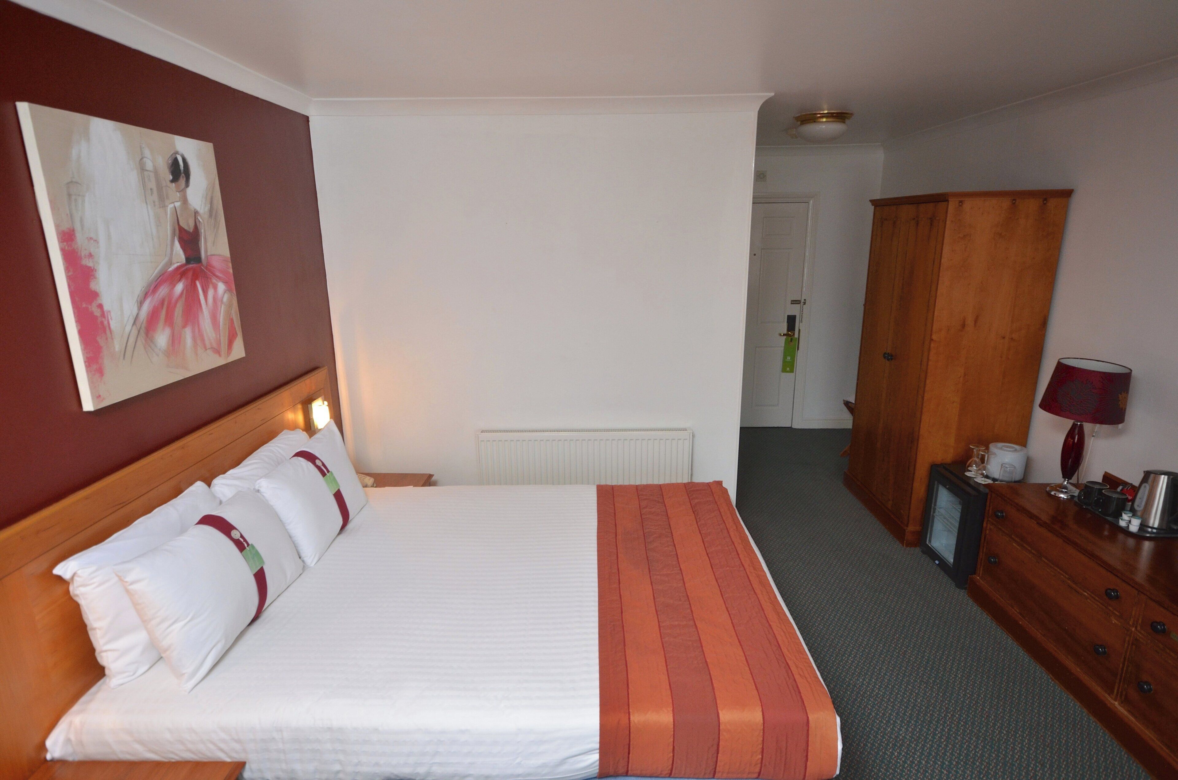 A hotel room with a bed, a wooden wardrobe, a desk with a lamp, and a painting on the wall.