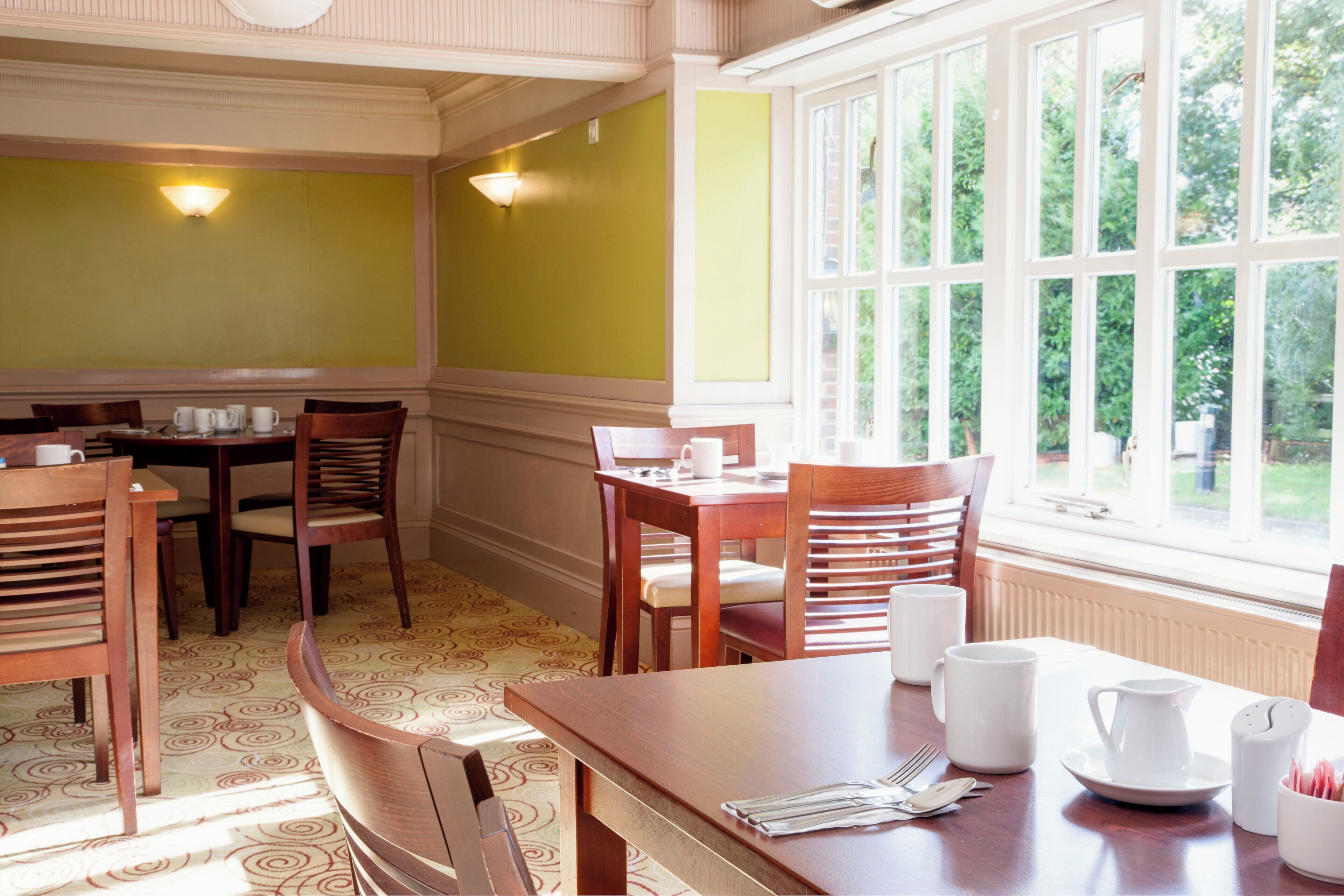 A dining area with wooden tables and chairs, a window with a view of greenery, and wall-mounted light fixtures.