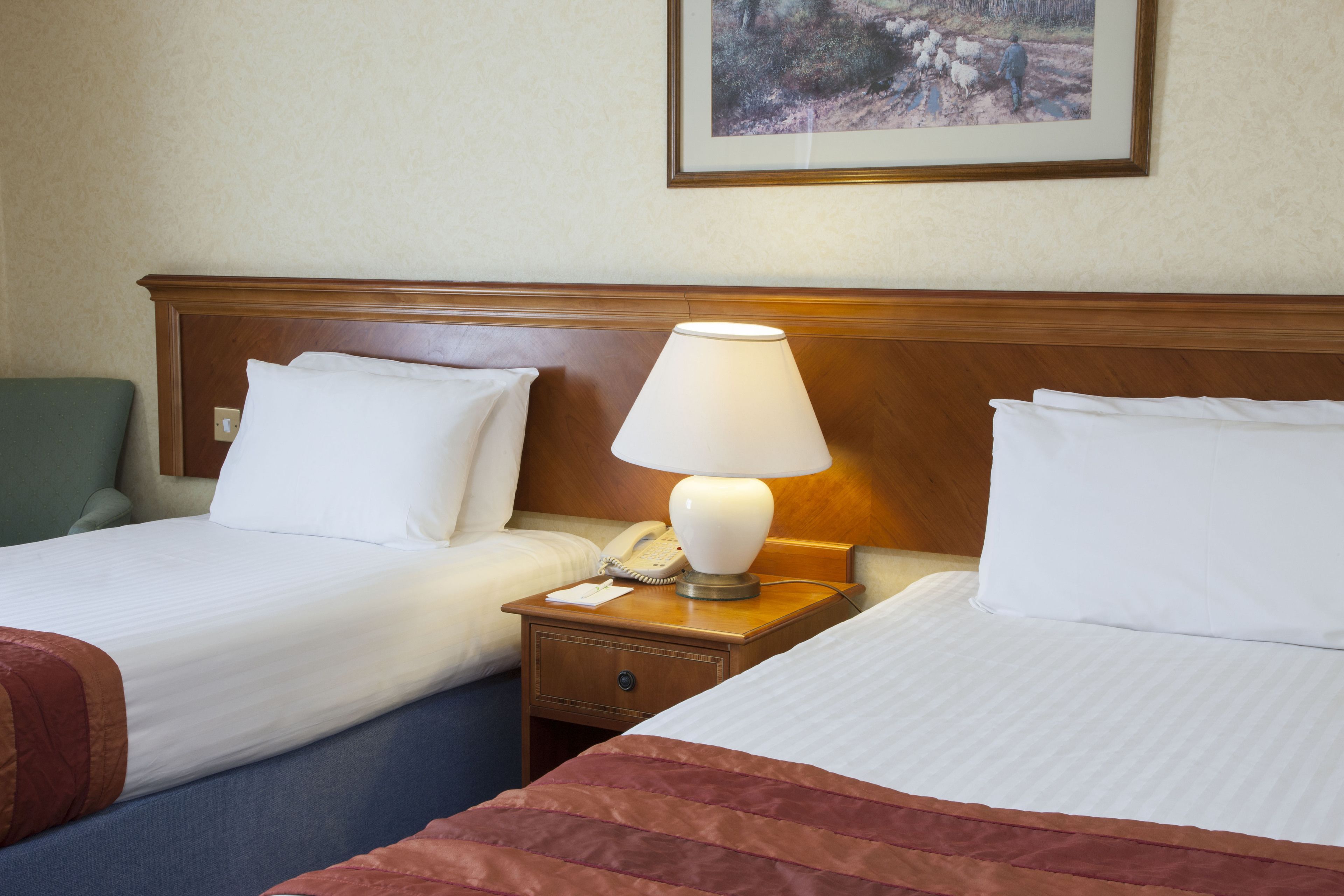 Ein Hotelzimmer mit zwei Betten, jeweils mit weißer Bettwäsche und einem h ölzernen Kopfteil. Dazwischen steht ein Nachttisch mit einer Lampe und einem Telefon.