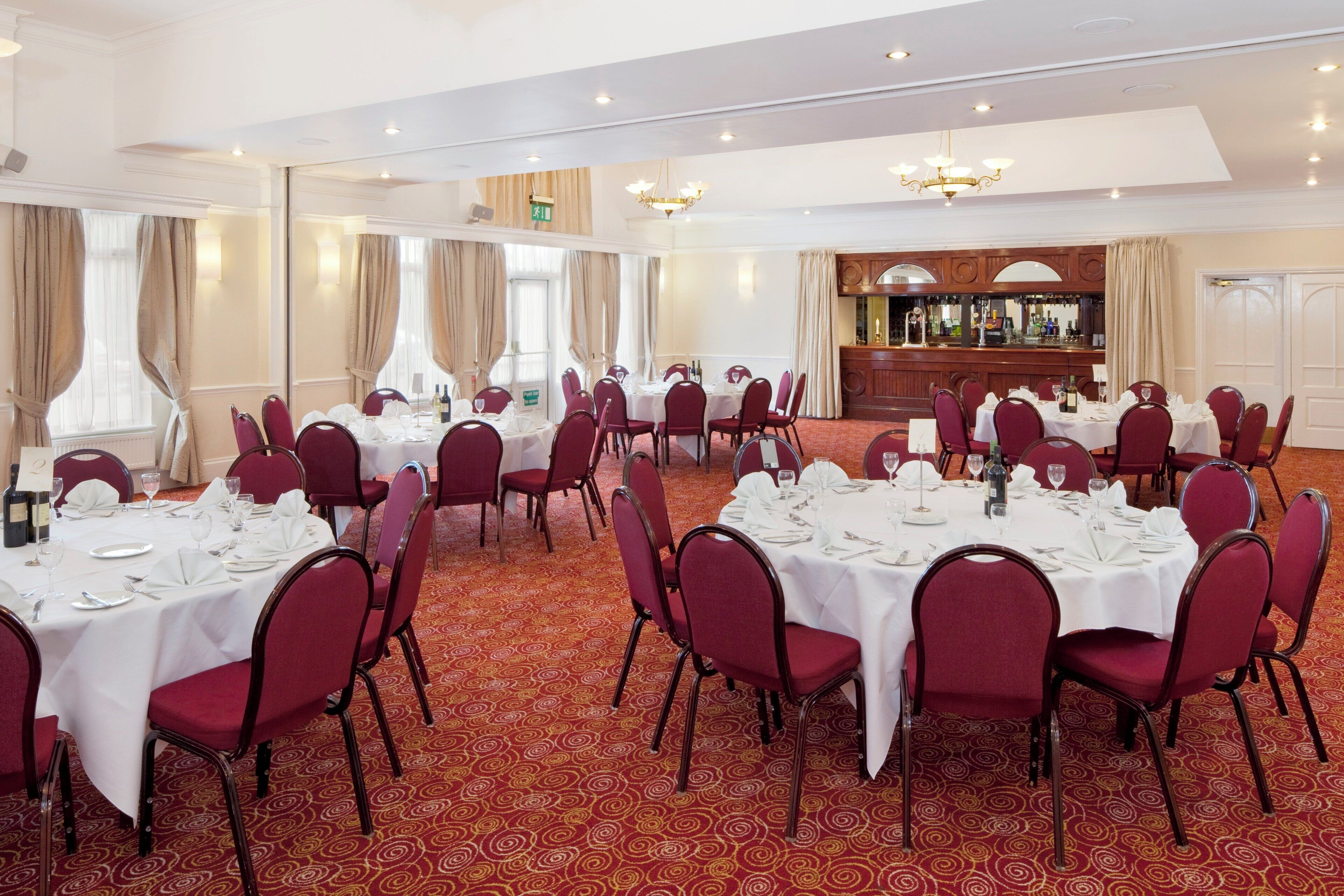 A banquet hall with round tables set for dining, red upholstered chairs, and a bar area.