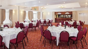 A banquet hall with round tables set for dining, red upholstered chairs, and a bar area.