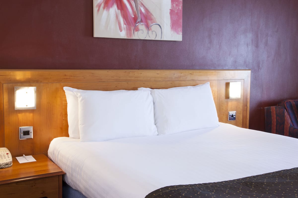 A hotel room with a wooden headboard, white bedding, a bedside table with a telephone, and a wall-mounted light.