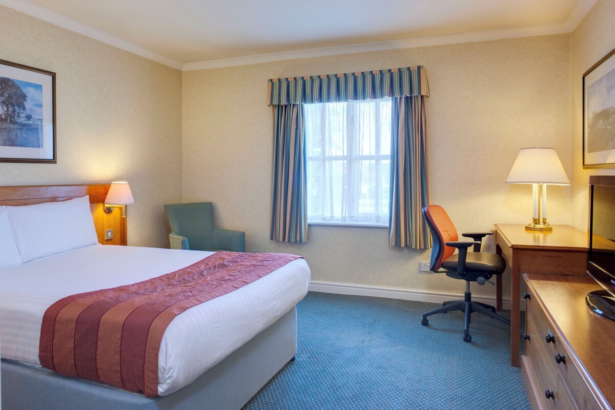 A hotel room with a bed, a desk, a chair, a lamp, and a window with curtains.