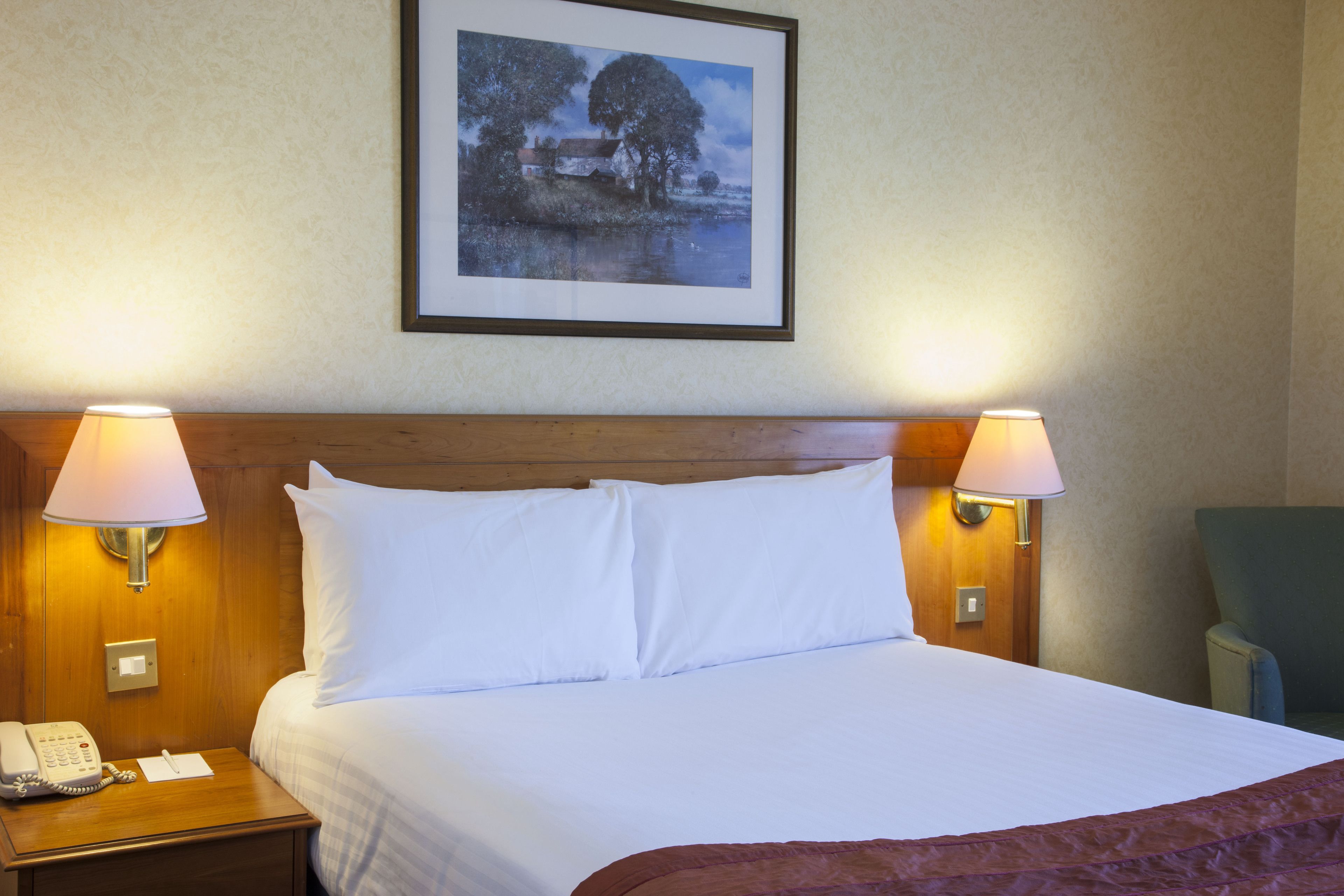 Hotelkamer met groot bed, twee bedlampen, een ingelijst schilderij aan de muur, een houten hoofdbord, een nachtkastje met telefoon en notitieboekje, en een groene stoel.