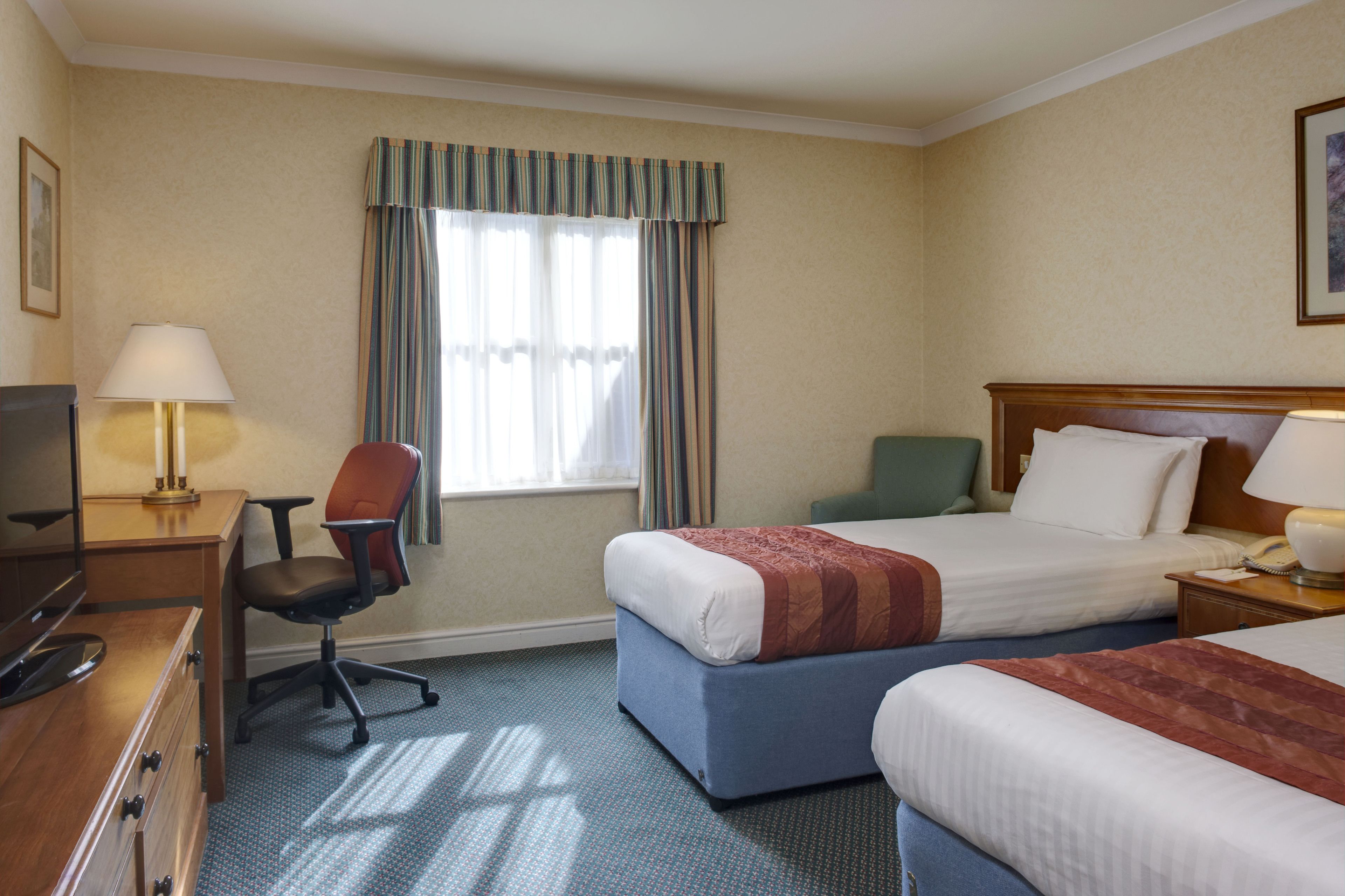 A hotel room with two beds, a desk with a lamp, a chair, and a window with curtains.