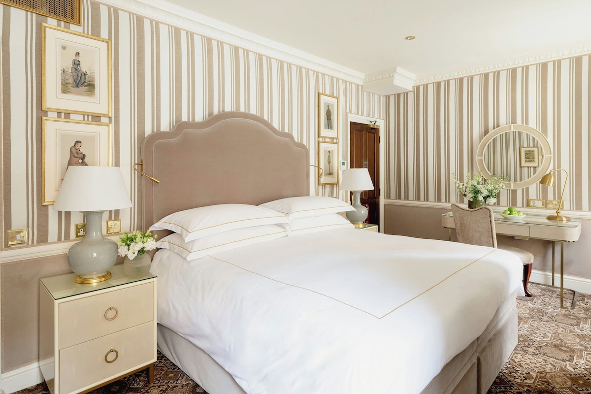 A neatly arranged hotel room with a large bed, a bedside table with a lamp, a desk with a chair, and a mirror.