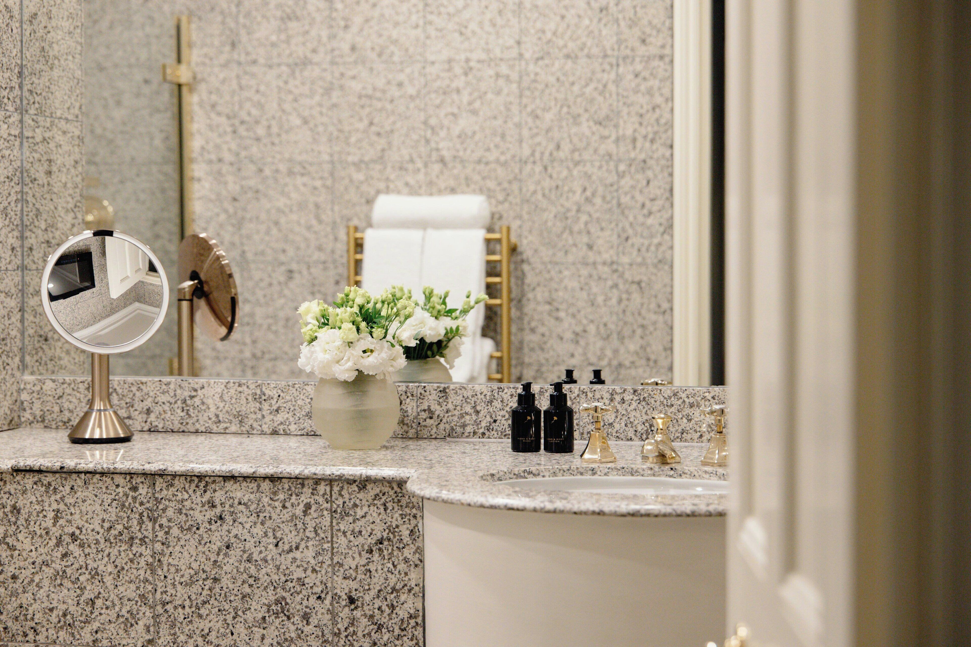 A bathroom with a granite countertop, a round mirror, a vase of flowers, and a towel rack with a white towel.