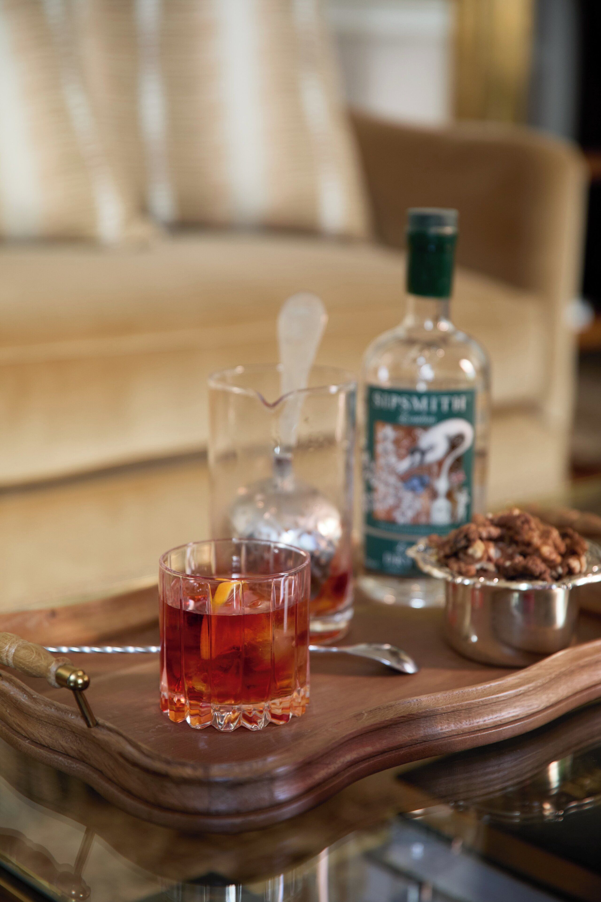 Uma bandeja com um copo de líquido âmbar, uma garrafa de licor e uma tigela de metal com nozes sobre uma mesa de madeira.
