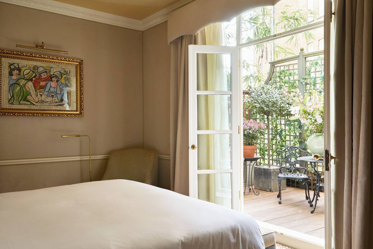 A bedroom with a large bed, a painting on the wall, and an open door leading to a balcony with plants and outdoor furniture.