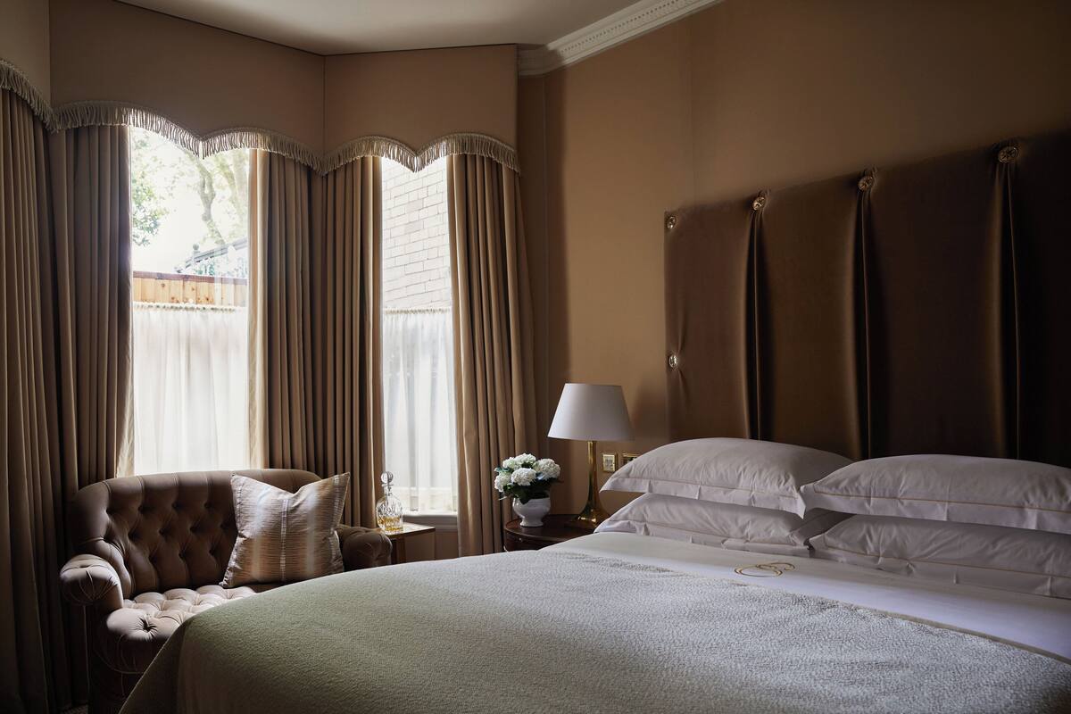 A hotel room with a large bed, a sitting area with a chair, a nightstand with a lamp, and curtains.