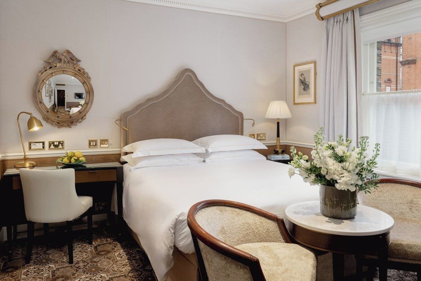 A hotel room with a large bed, a desk with a lamp, a chair, a small table with a vase of flowers, and a mirror on the wall.