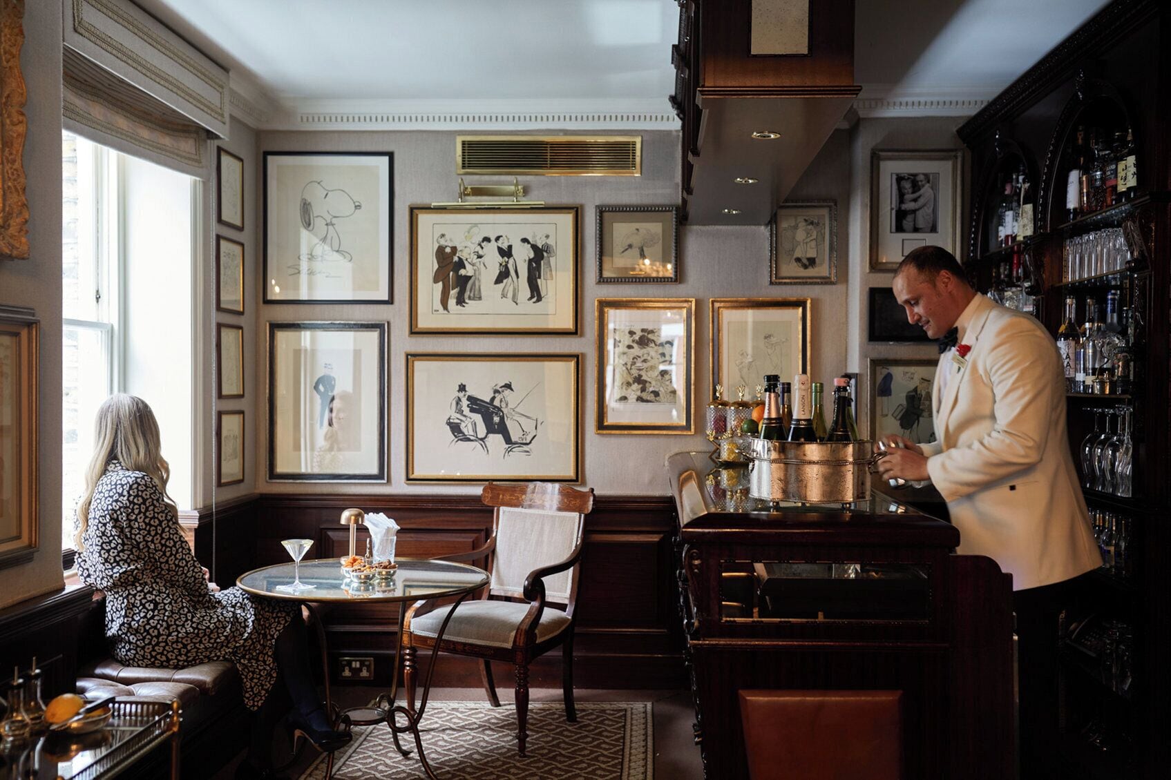 A bar area with a bartender, a table with a glass and a bottle, and framed artwork on the walls.