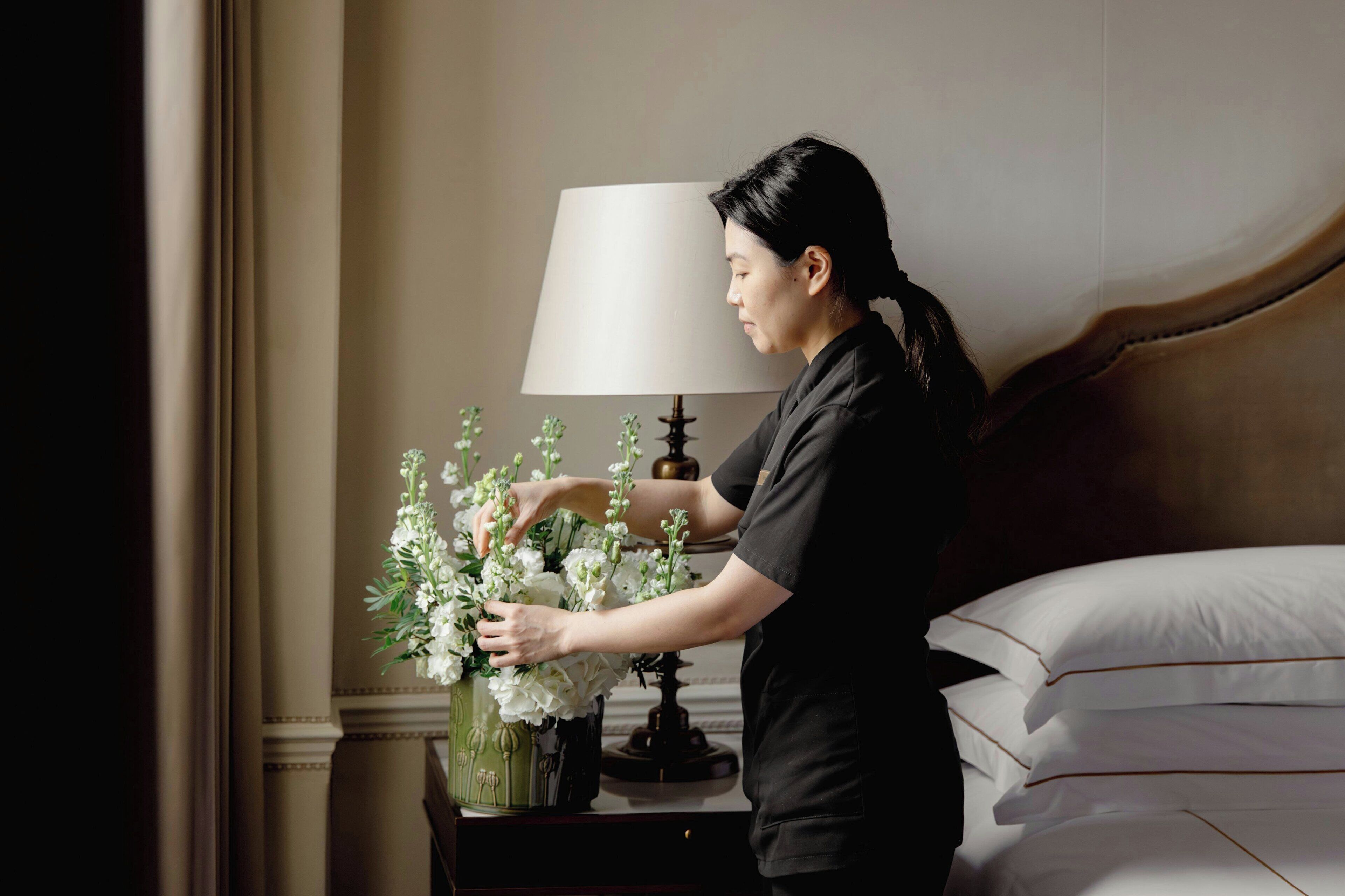 En person som arrangerar vita blommor i en vas på ett nattduksbord bredvid en lampa i ett hotellrum.