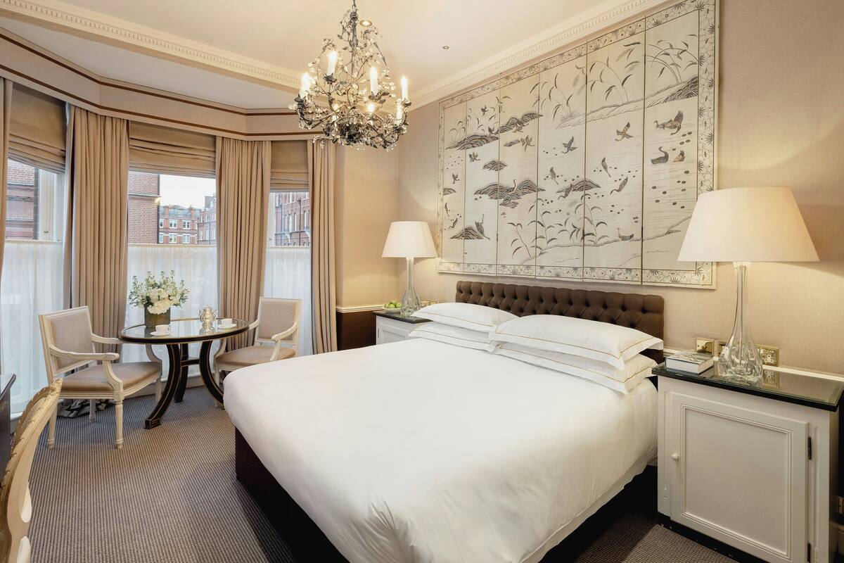 A hotel room with a large bed, two bedside tables, a chandelier, a wall with a nature-themed artwork, a round table with chairs, and a view of buildings through large windows.