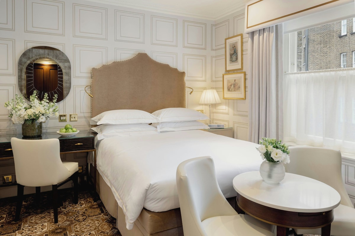A hotel room with a large bed, two chairs, a small table, a mirror, and a window with curtains.
