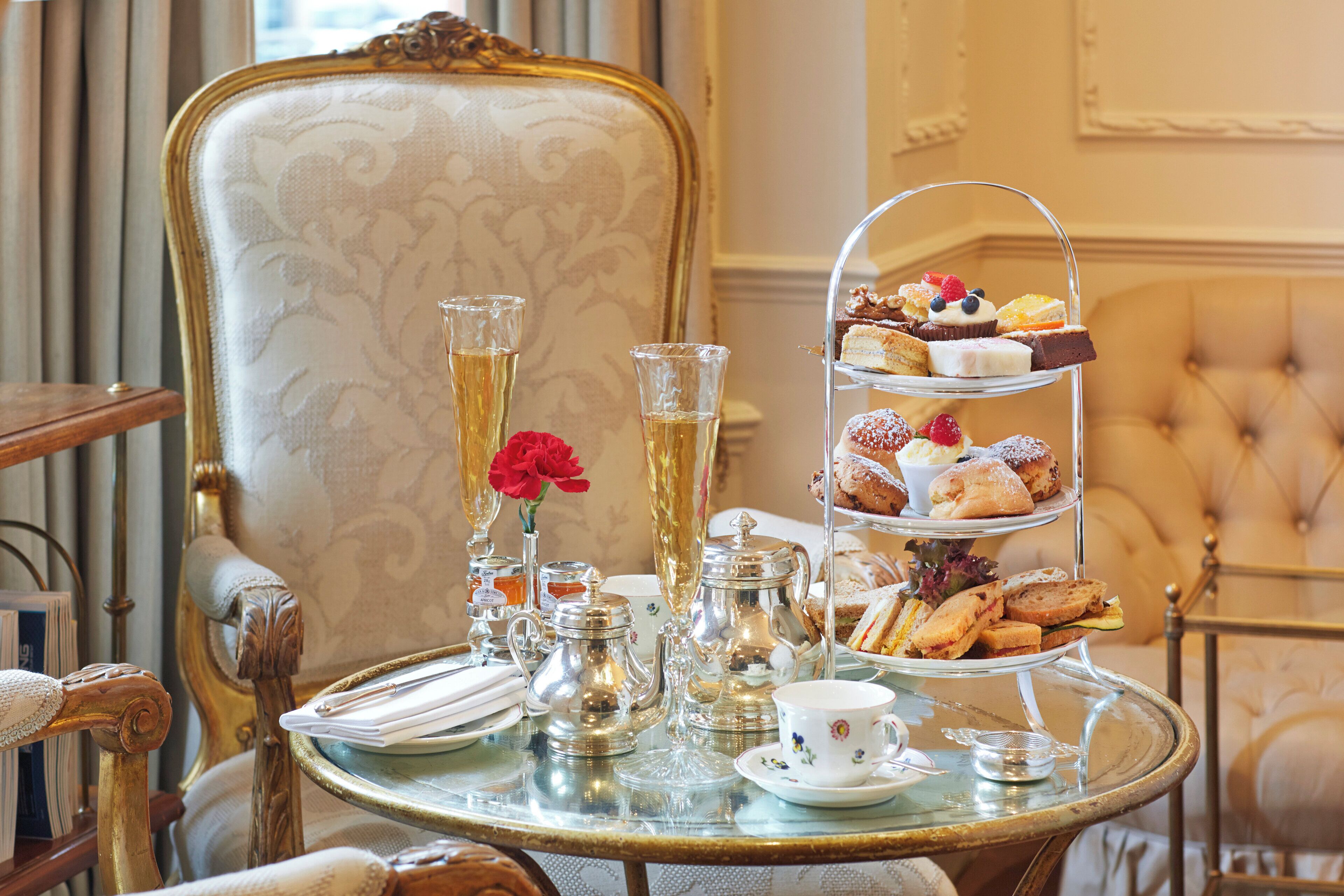 A three-tiered dessert stand with various pastries and a glass of a golden beverage, a tea set, and a vase with a red flower on a glass table.