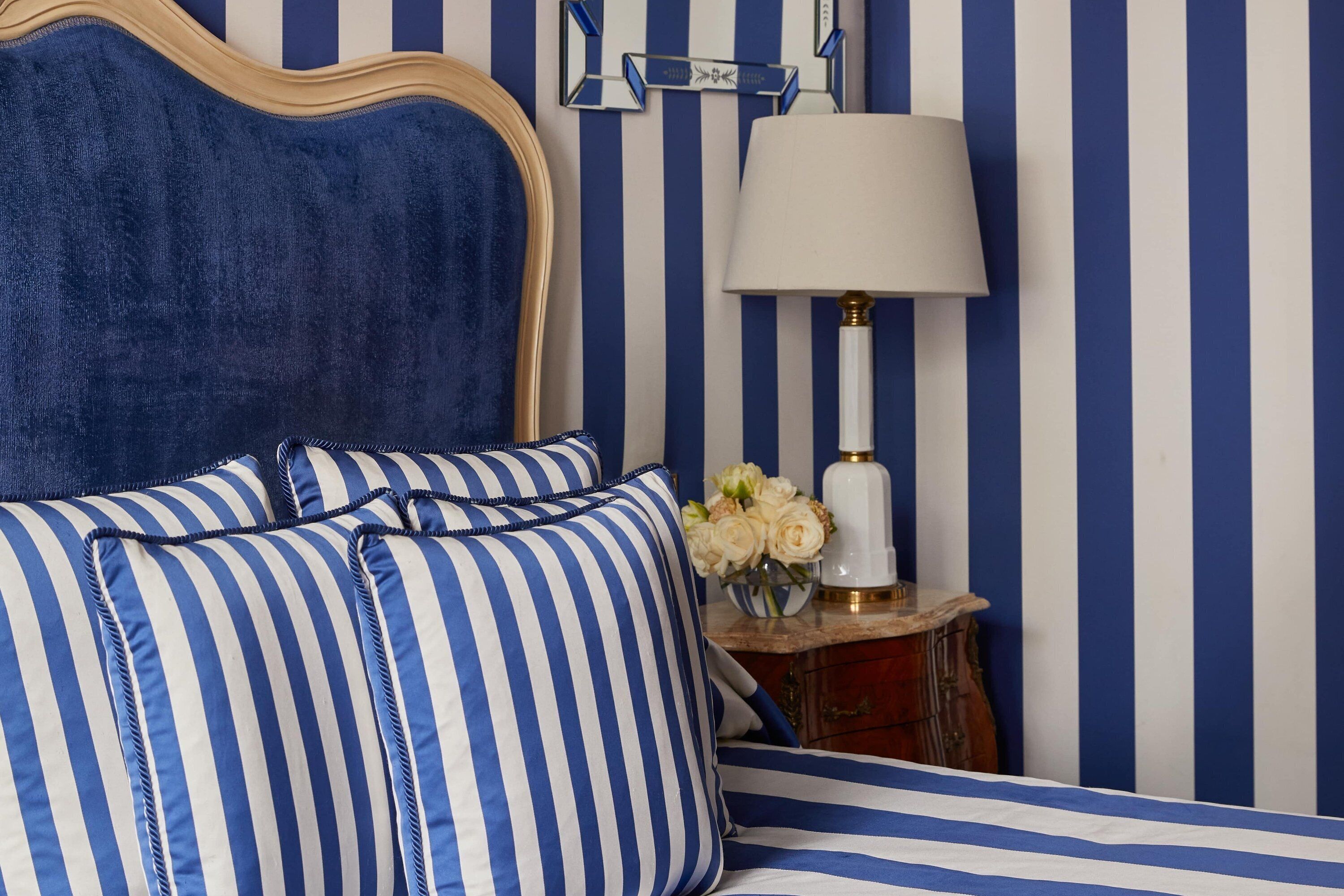 Uma cama com roupa de cama listrada em azul e branco, uma cabeceira de madeira e uma mesa de cabeceira com abajur e flores.