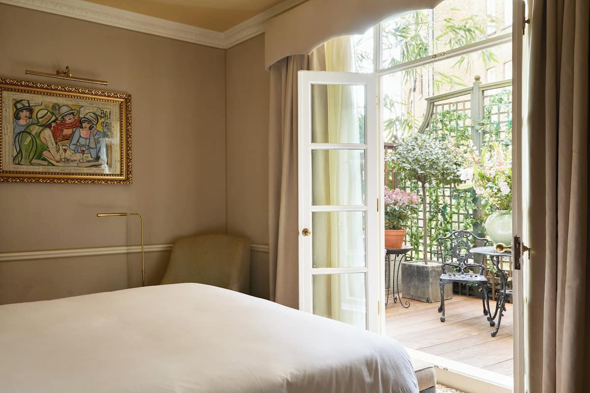 A bedroom with a large bed, a painting on the wall, and an open door leading to a balcony with plants and outdoor furniture.