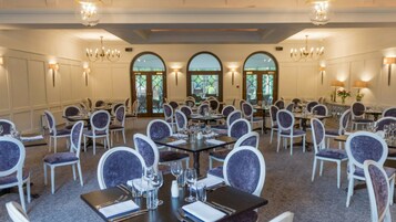 Uma sala de banquetes com mesas redondas, cadeiras, lustres e grandes janelas.