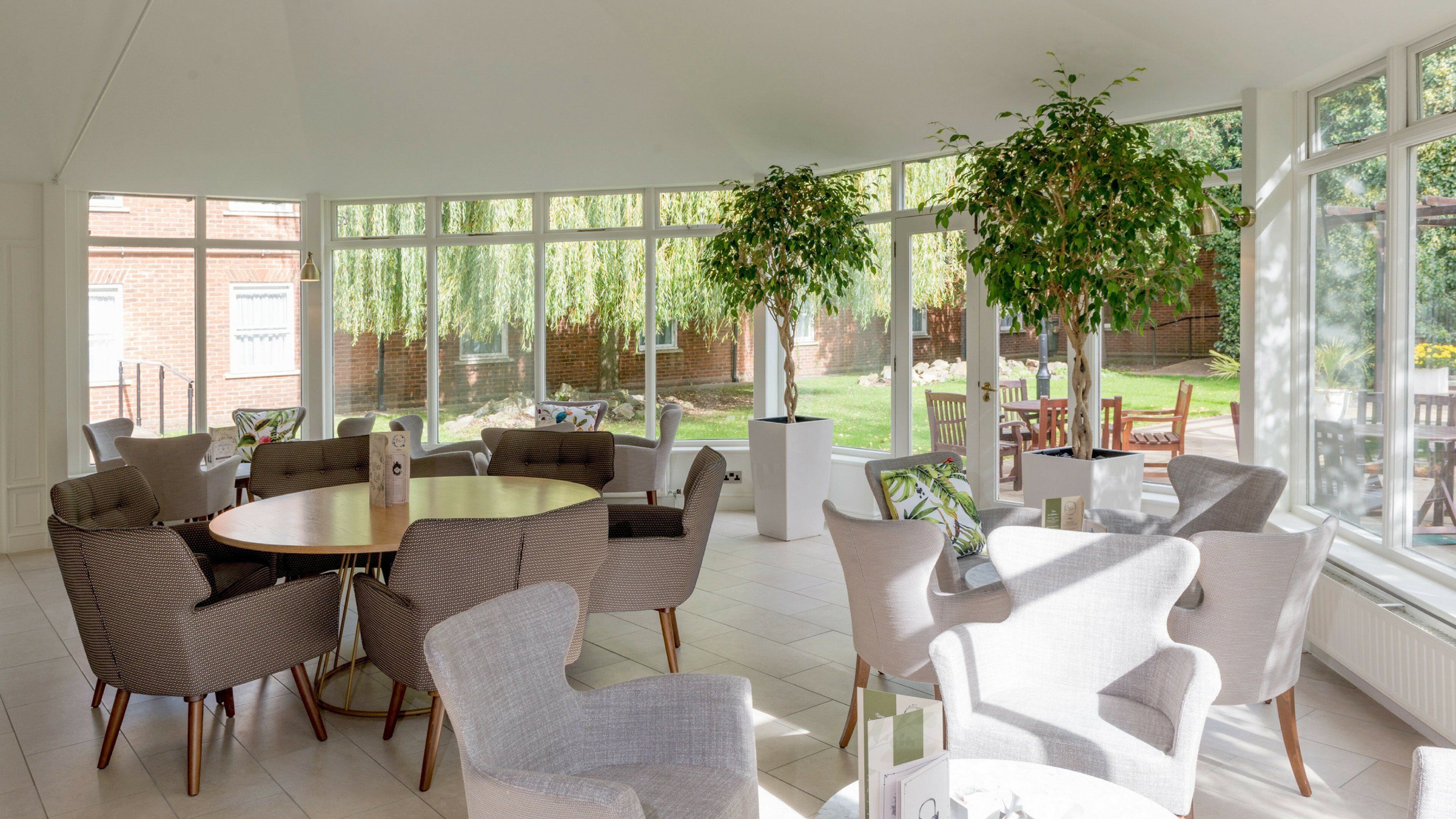 Une pièce spacieuse avec une table ronde et plusieurs chaises, de grandes fenêtres et des plantes d’intérieur.