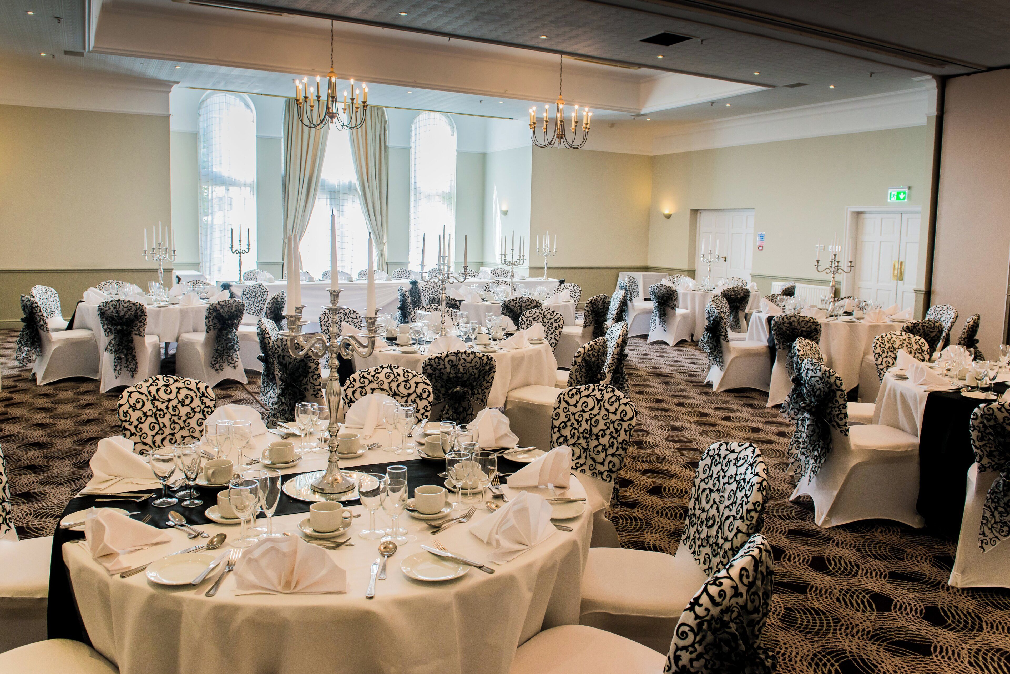 Un salón de banquetes con mesas redondas dispuestas para un evento formal, con manteles blancos y cubre-sillas estampados negros.