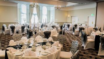 Un salón de banquetes con mesas redondas dispuestas para un evento formal, con manteles blancos y cubre-sillas estampados negros.